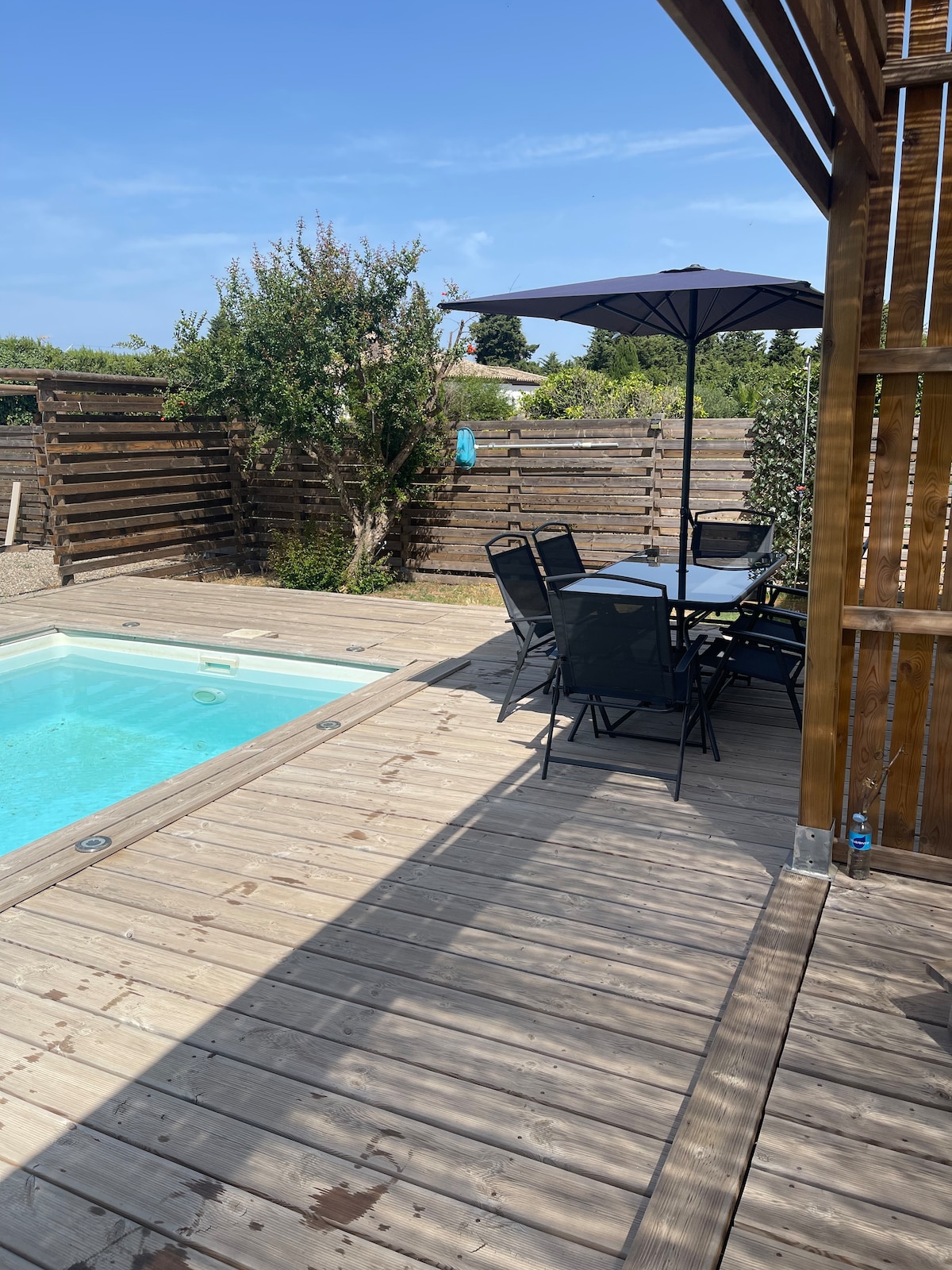 A wooden deck area is shown with a clear swimming pool and a rectangular dining table surrounded by six black chairs. A large umbrella provides shade, while greenery borders the space, creating a sense of privacy and relaxation.