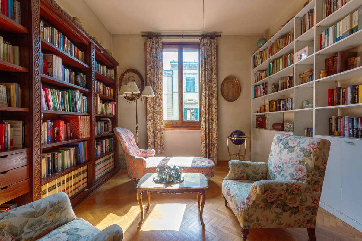 A comfortable sitting area is surrounded by two wooden bookcases filled with books. Two floral armchairs are placed opposite a small coffee table, while natural light enters through a large window adorned with patterned curtains. A wall-mounted clock and decorative globe add character to the space.