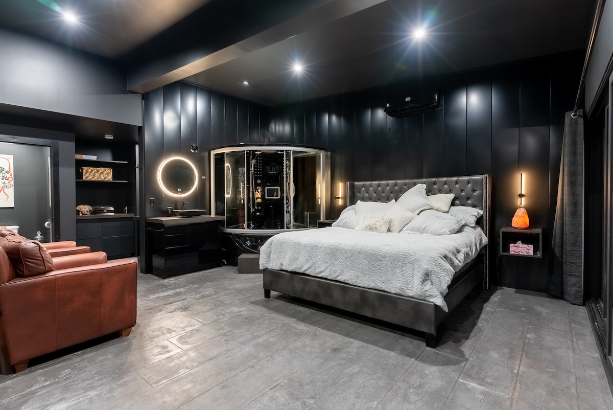 The spacious master bedroom features a large king-size bed with soft bedding and a contrasting dark colour palette. A relaxation area with cozy leather chairs and soft lighting complements the luxurious steam capsule and rain shower enclosure, creating a serene atmosphere.