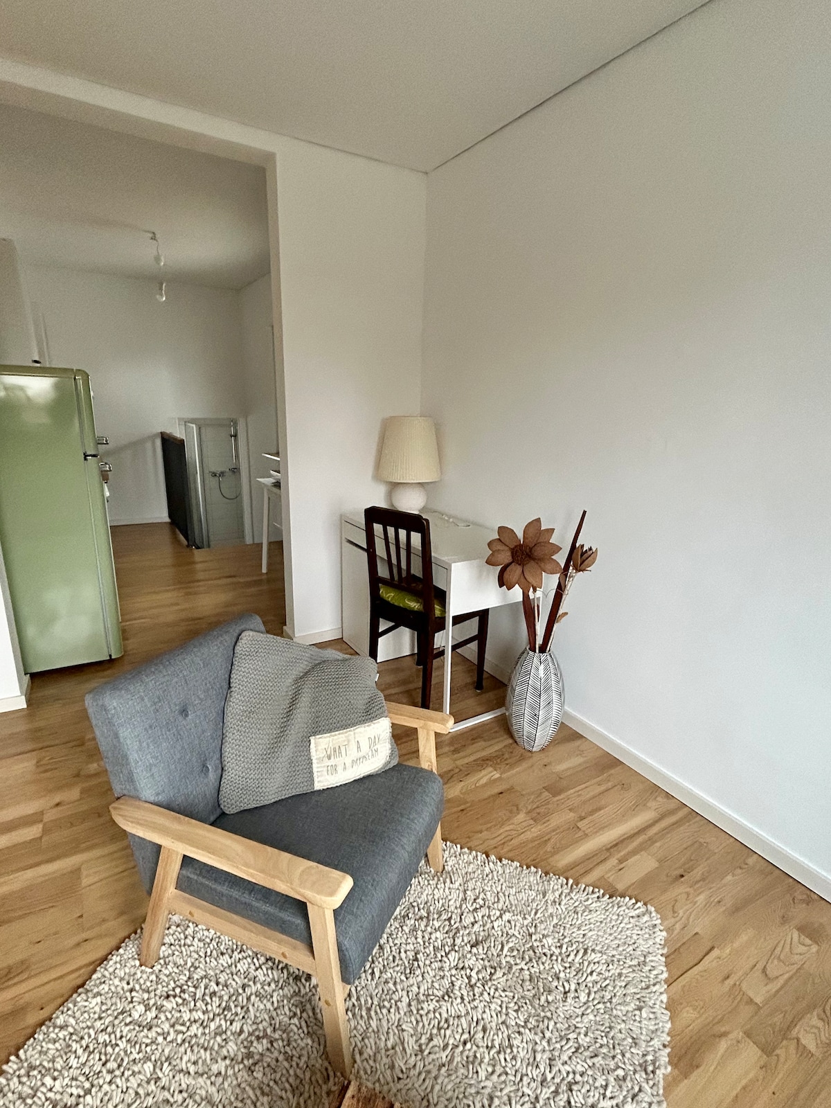 A cozy seating area features a soft gray armchair placed on a pale rug. A small desk with a lamp and a decorative vase containing dried plants is visible. Light oak flooring extends throughout the space, complemented by a green vintage refrigerator in the background.