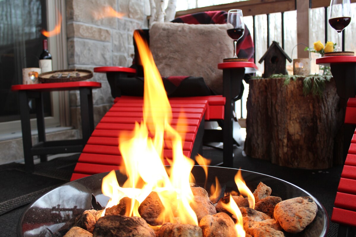 A fire pit with glowing flames flickers, surrounded by a circular arrangement of smooth stones. Red lounge chairs are positioned nearby on a covered porch, accompanied by glasses of red wine on small tables. Decorative elements include a tree stump and a small birdhouse.