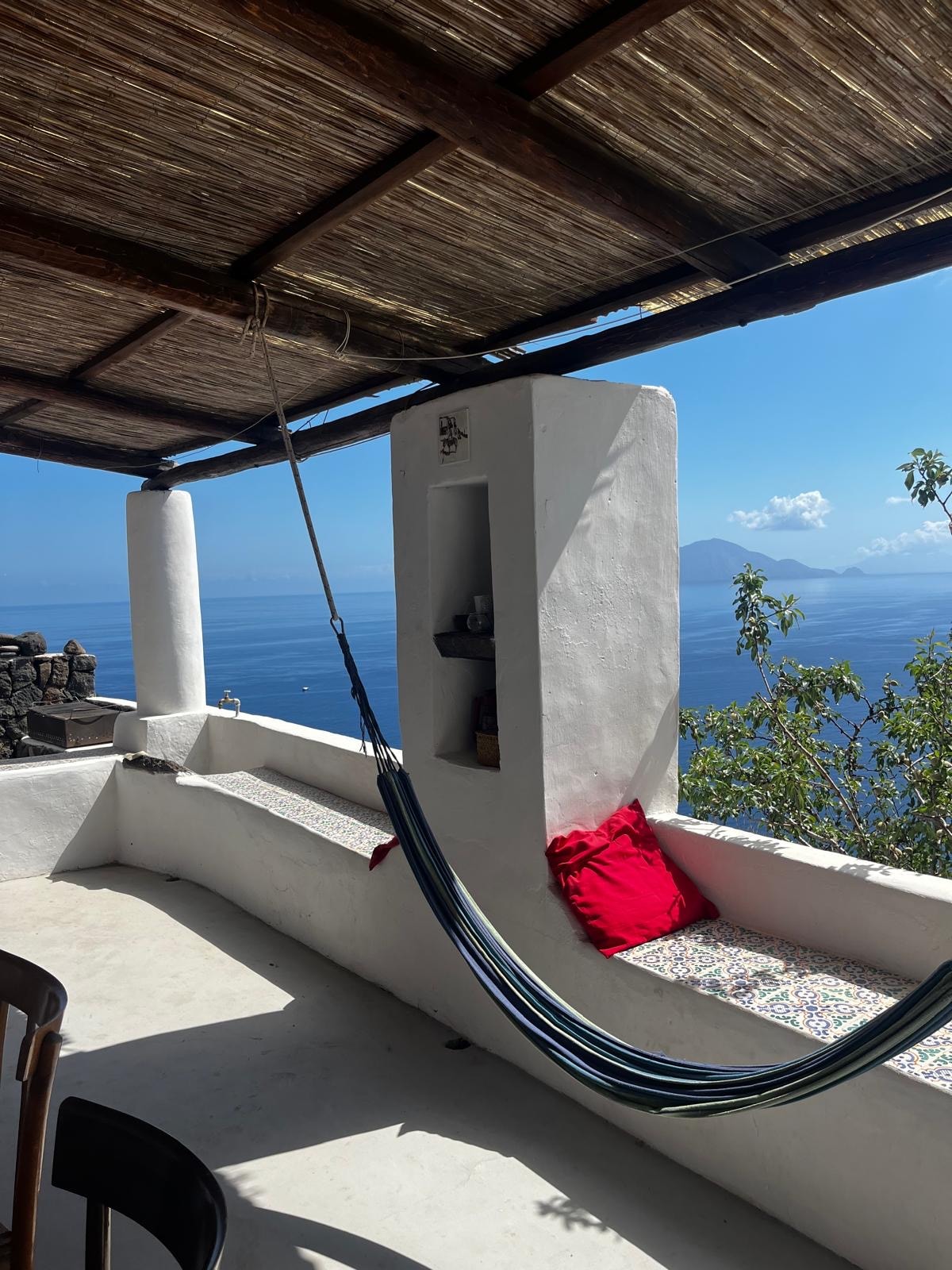 A shaded terrace offers a serene space with a hammock hanging freely. A cozy red cushion is placed on the built-in bench, complemented by white walls and a view of the azure sea. Natural light brightens the area, highlighting the peaceful ambiance.