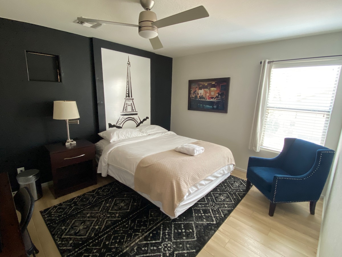 A spacious bedroom features a bright ceiling fan above the bed, adorned with light linen. A stylish accent wall showcases an Eiffel Tower painting. A comfortable blue chair complements a dark wood nightstand, while natural light filters through the window, highlighting the laminate wood flooring.
