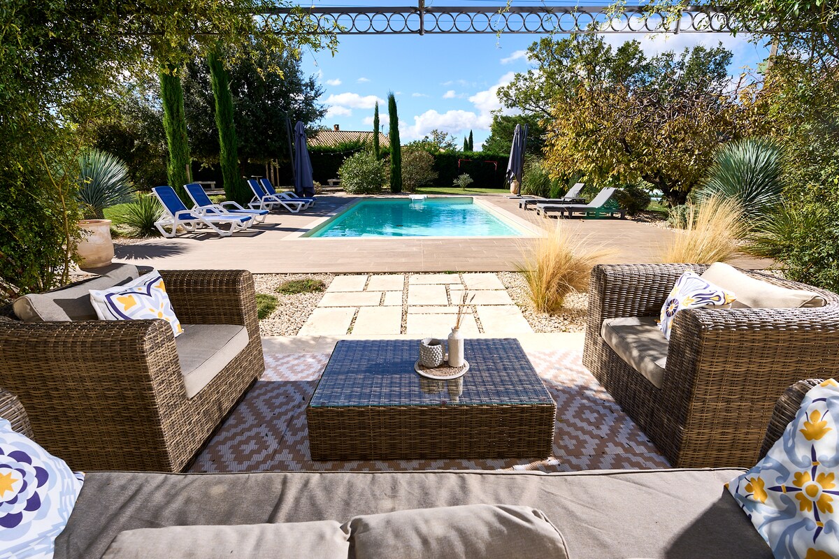 An outdoor lounge area features two wicker chairs and a glass-topped table, positioned to overlook a private swimming pool. Surrounding lush greenery and well-maintained landscaping are visible, along with sun loungers placed near the pool, creating a serene and inviting atmosphere.