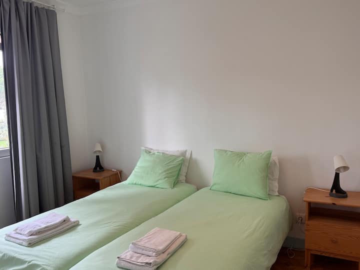 Quarto com duas camas individuais, roupa de cama clara e luz natural. Ambiente simples, acolhedor e funcional, com mesas de apoio e decoração neutra para um descanso confortável.