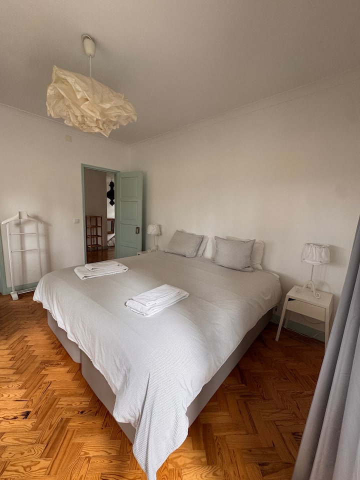 Quarto amplo e luminoso com cama king-size (1.80x2.00m), decoração minimalista e chão de madeira. A luz natural cria um ambiente acolhedor e tranquilo, ideal para descanso.