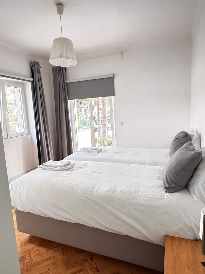 Quarto luminoso com duas camas individuais juntas, roupa de cama branca e janelão com cortinas cinzentas. Ambiente acolhedor, ideal para descanso tranquilo com vista e luz natural do jardim.
