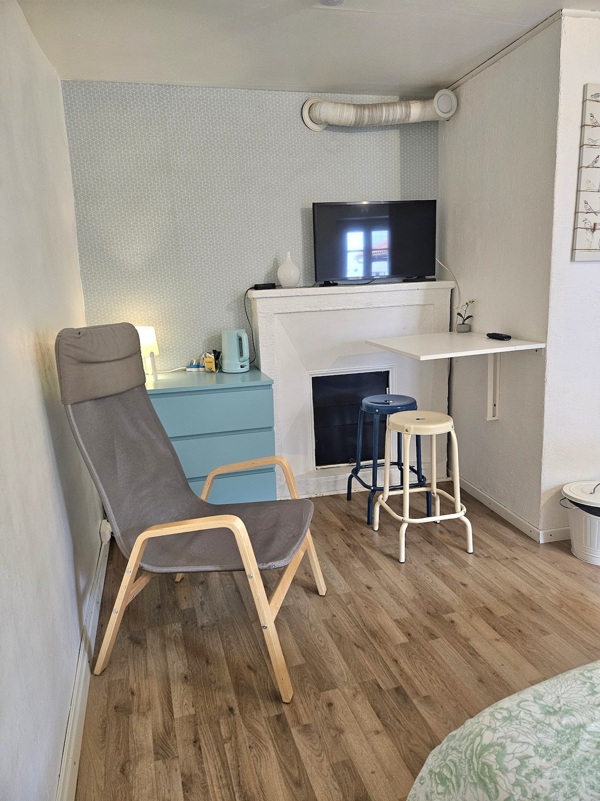A compact corner of the studio is presented, showcasing a two-toned grey chair beside a light blue cabinet. A flat-screen television is mounted above a minimalist fireplace, complemented by a small table with two stools. Natural light filters through a window, enhancing the functional space.