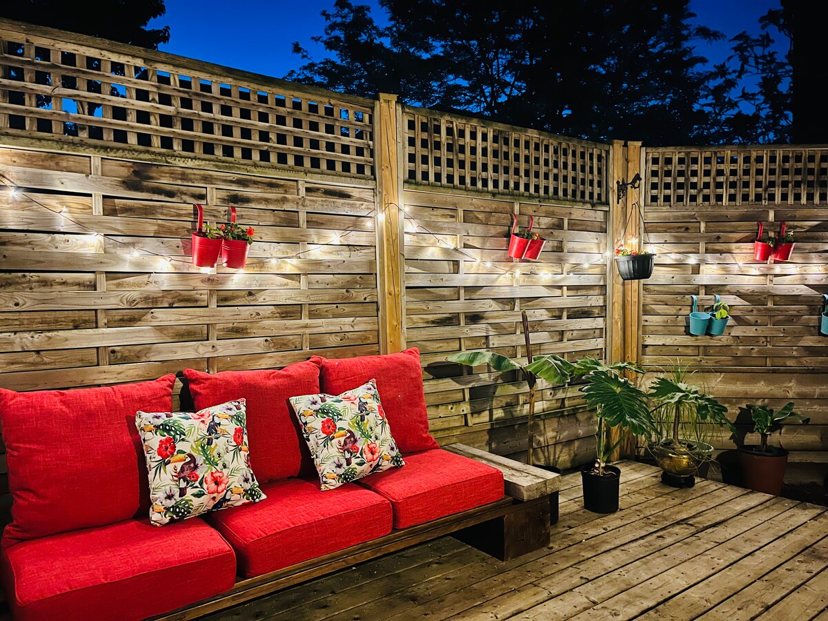 A cozy terrace area features a red couch adorned with floral pillows, positioned against a wooden privacy fence. Planters hang on the wall, showcasing vibrant greenery, while warm string lights create a soft ambiance, enhancing the inviting outdoor space.