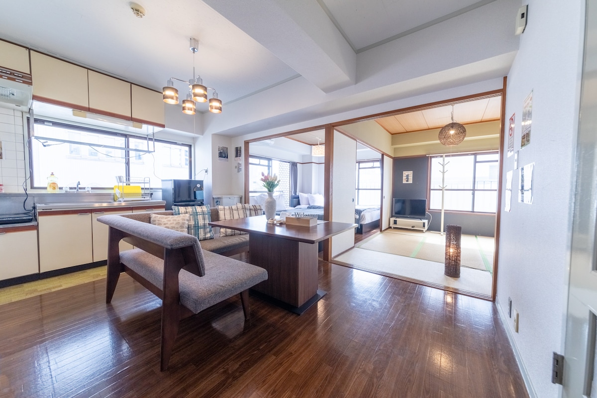 The open-plan living area features a dining table with a wooden surface and benches, surrounded by large windows allowing natural light. A seamless transition is observed to the tatami room on one side and a cozy lounge area on the other, creating a spacious atmosphere.