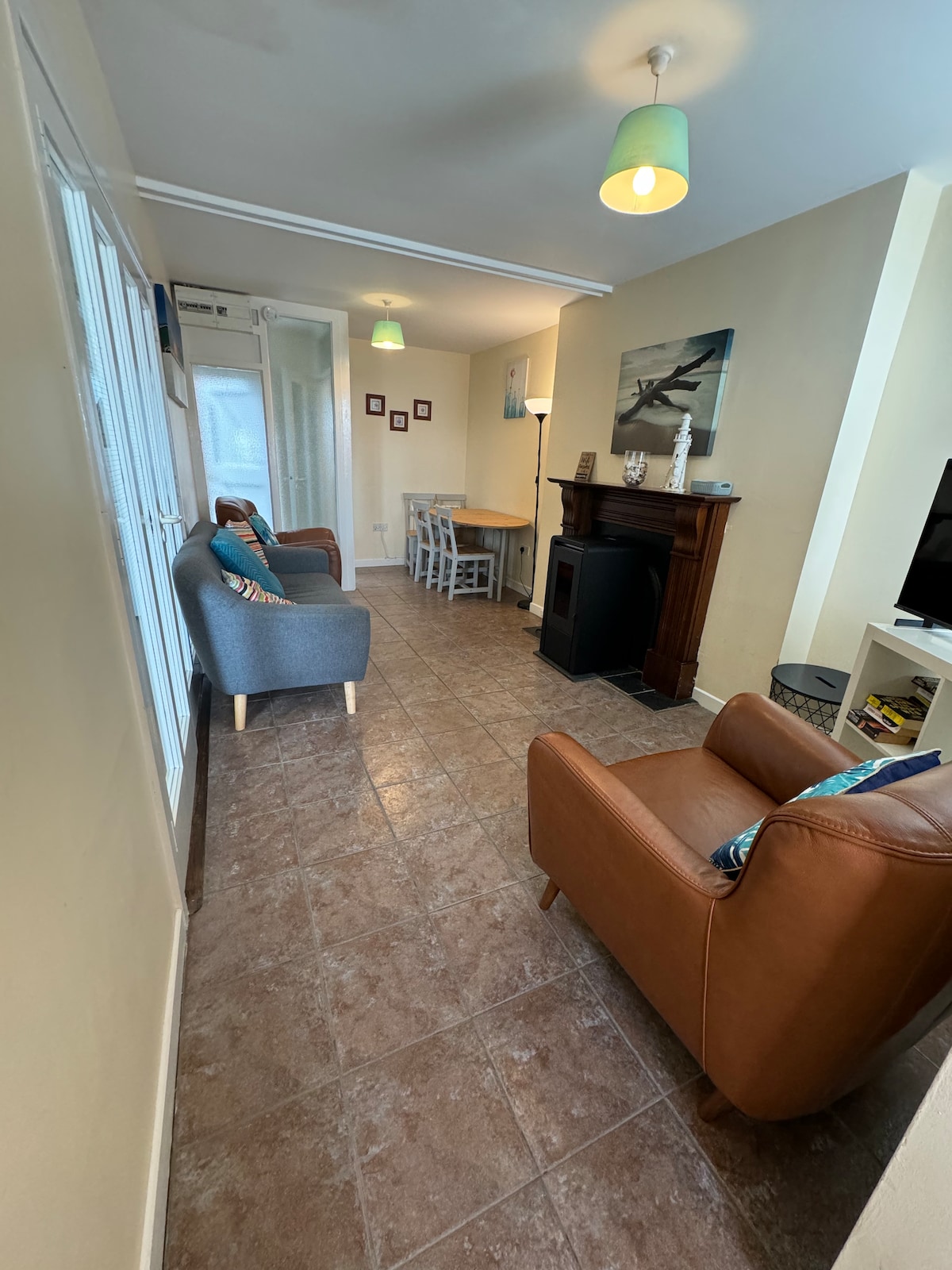 The open plan living area contains a mix of seating options, including two armchairs and a small dining table with chairs. A wood pellet stove is positioned near the seating, contributing to the cozy atmosphere. Light from the overhead fixtures illuminates the neutral-colored walls.
