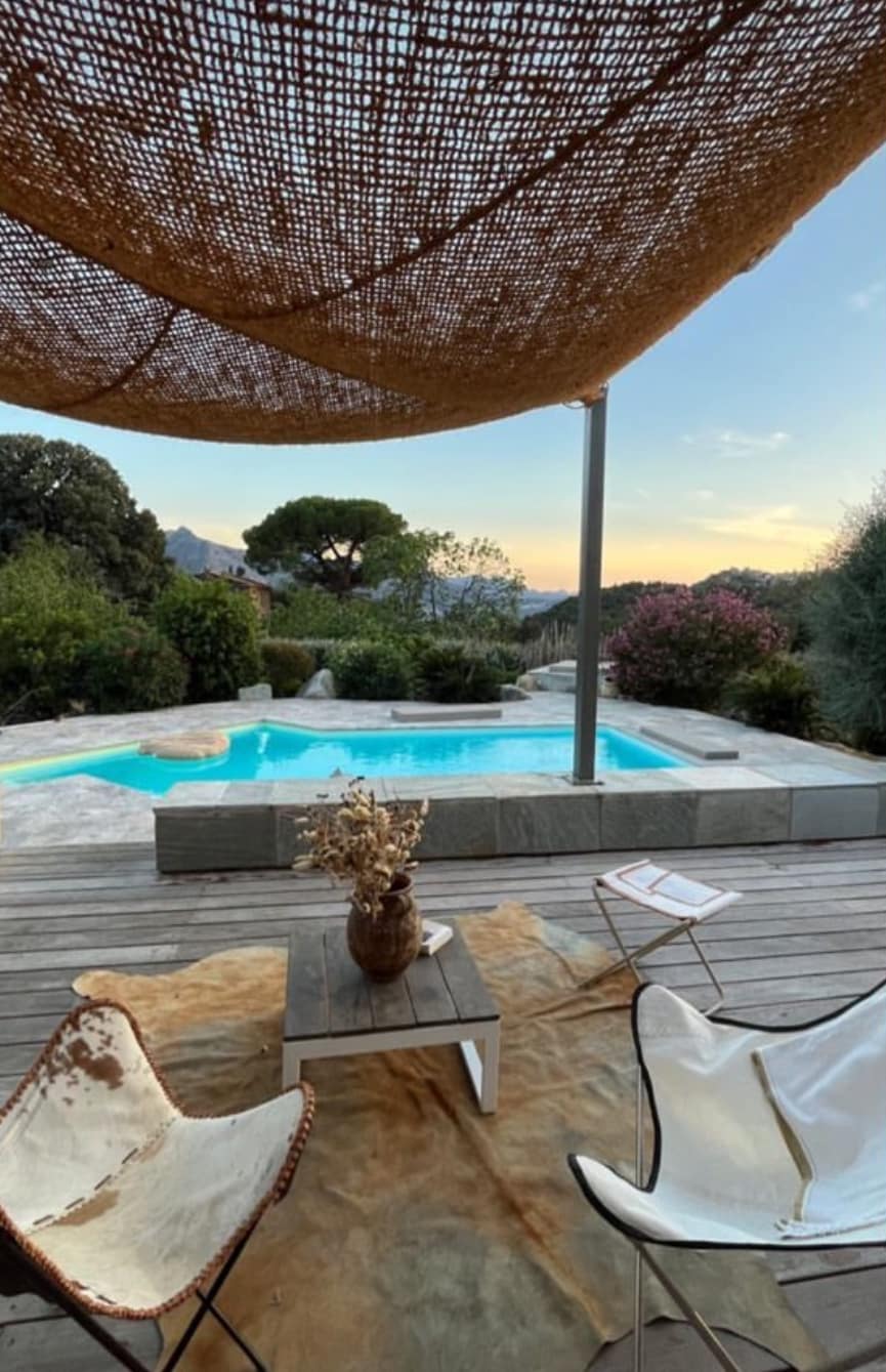 An outdoor lounging area features two white chairs with simple design, positioned around a rustic wooden table. A shaded area is created by a woven canopy above, while a swimming pool with light blue water is visible nearby, surrounded by lush greenery and ornamental plants.