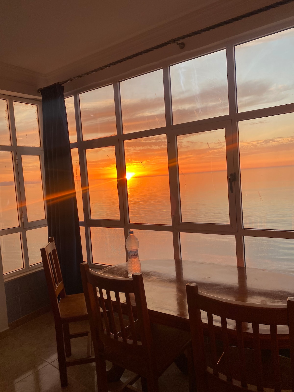 A dining area is illuminated by a warm sunset casting golden hues through large windows. A wooden table is accompanied by four chairs, while a water bottle rests on the table's surface. The sea is visible beyond the glass, creating a serene atmosphere.