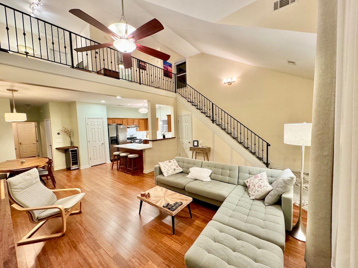 An open living area features a spacious sectional sofa and a light wood coffee table, complemented by modern wooden chairs. The vaulted ceiling enhances the sense of space, while large windows allow natural light to illuminate the room. A dining area and kitchen are visible in the background.