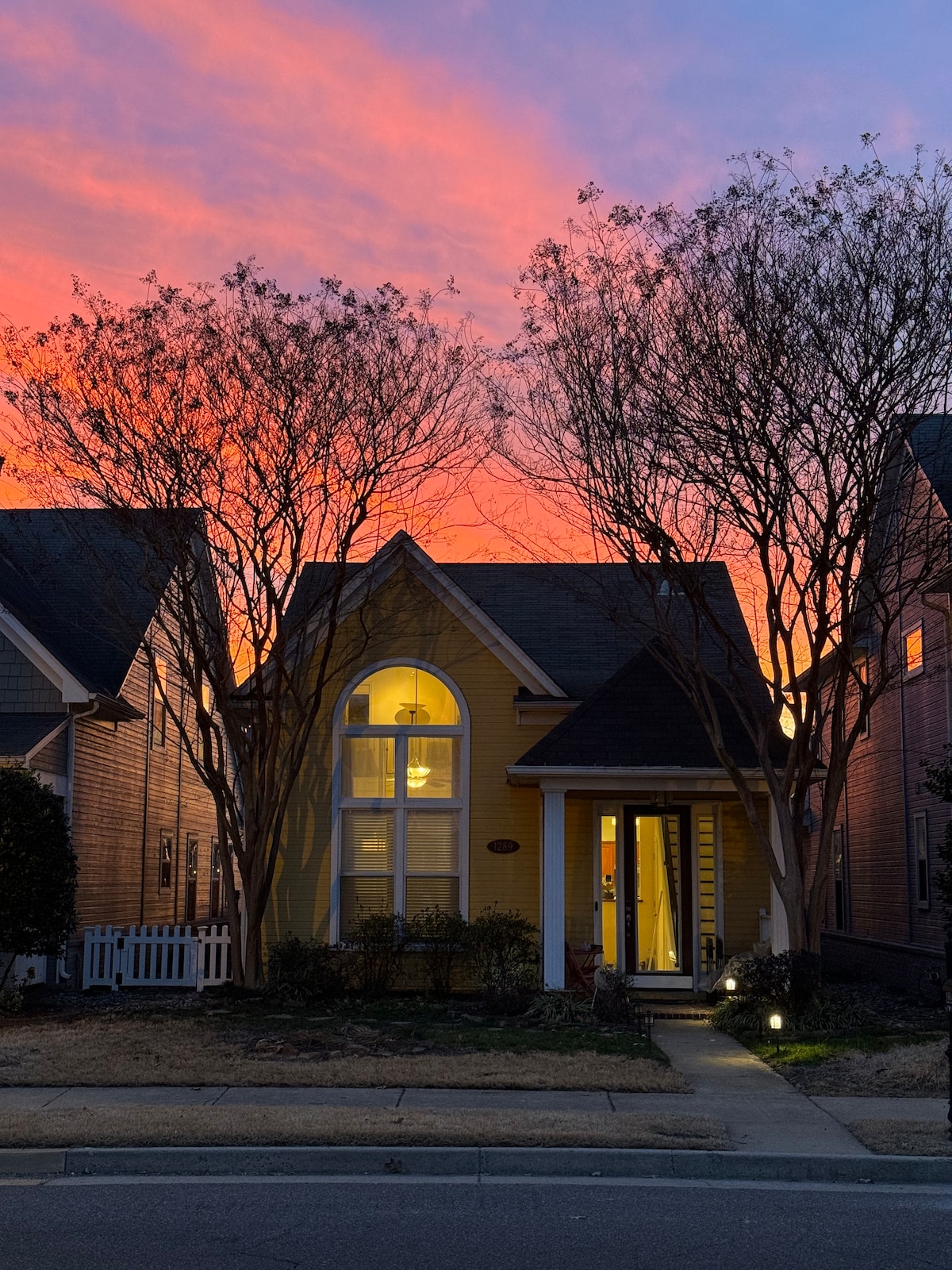 The exterior of a charming yellow house is framed by two trees as the sky is painted in hues of pink and orange at sunset. Pathway lighting softly illuminates the entrance, which features an arched window above the door.