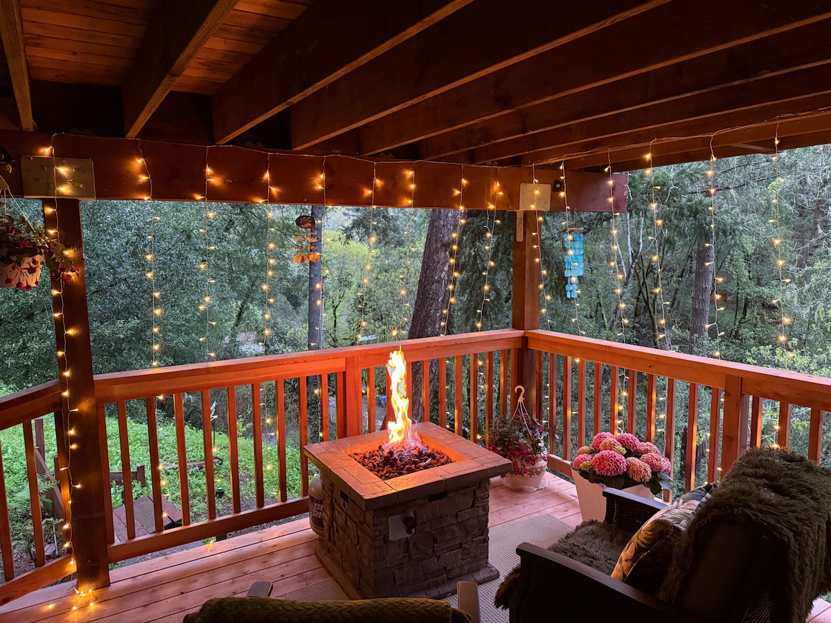 A private covered porch is illuminated by string lights, creating a serene ambiance. A stone fire pit centers the space, with flames dancing above. Comfortable seating with decorative pillows is available, surrounded by lush greenery and the sounds of nature.