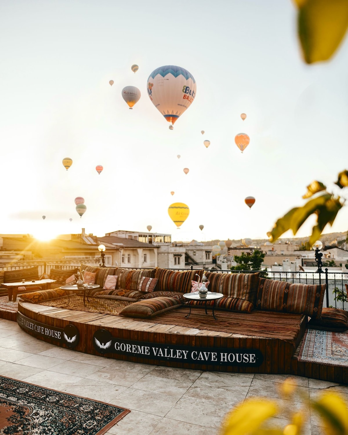 A spacious terrace area is adorned with traditional Turkish cushions and seating, inviting relaxation. Early morning sunlight illuminates the space while colorful hot air balloons drift in the sky above. The serene landscape of Göreme is visible in the background, enhancing the peaceful setting.