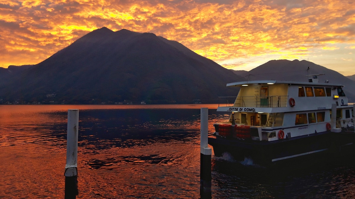 A ferry navigates the tranquil waters at sunset, with vibrant hues reflecting off the lake. The silhouette of a mountain rises in the background, creating an atmospheric scene that captures the beauty of the surrounding landscape.