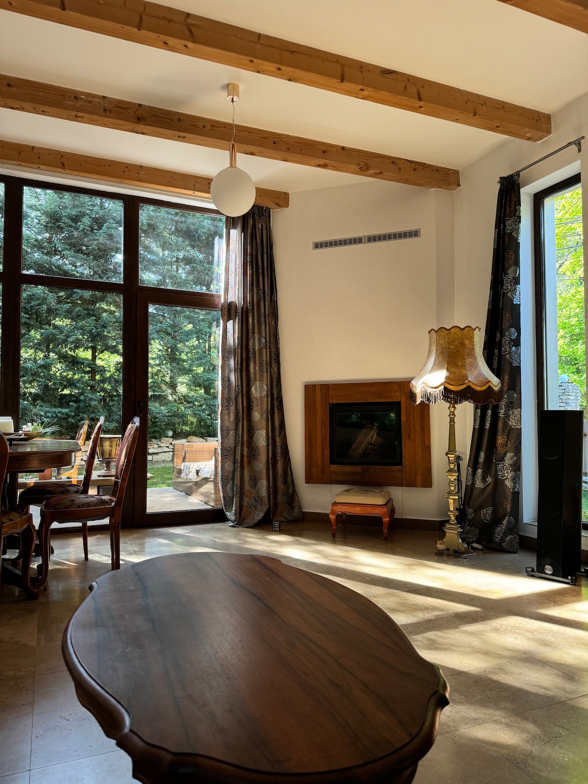A bright living area features large windows that overlook the lush outdoors. Elegant wooden beams are visible on the ceiling, while a stylish lamp and a coffee table anchor the space. A dining table and chairs are set nearby, creating an inviting setting.