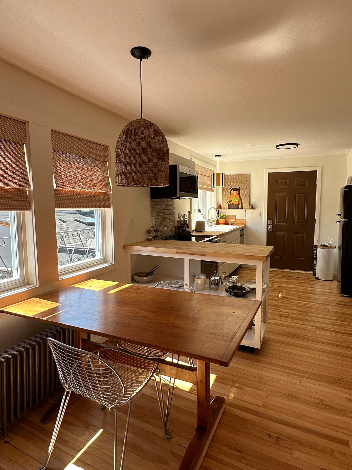 A bright kitchen area features a wooden dining table paired with a wire chair. Sunlight filters through the window treatments, illuminating the modern appliances and open shelving. Decorative elements, including artwork on the wall, enhance the welcoming environment.