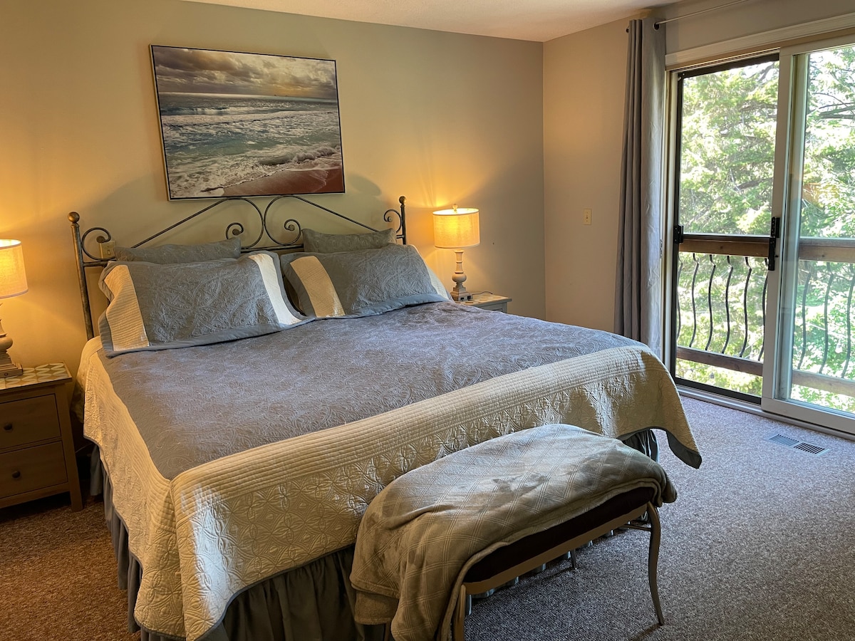 A spacious room features a large bed with decorative bedding, complemented by matching bedside lamps. Soft curtains frame the large glass door, allowing natural light to illuminate the space. A cozy bench sits at the foot of the bed, and trees are visible outside.