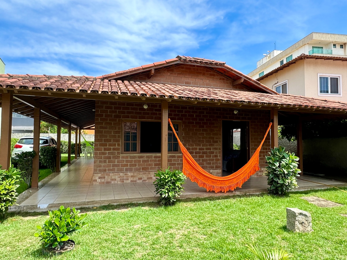The exterior view of the house features a spacious covered porch with a vibrant orange hammock, surrounded by manicured grass and small shrubbery. A tiled pathway leads to the entrance with large windows that allow natural light to fill the interior.
