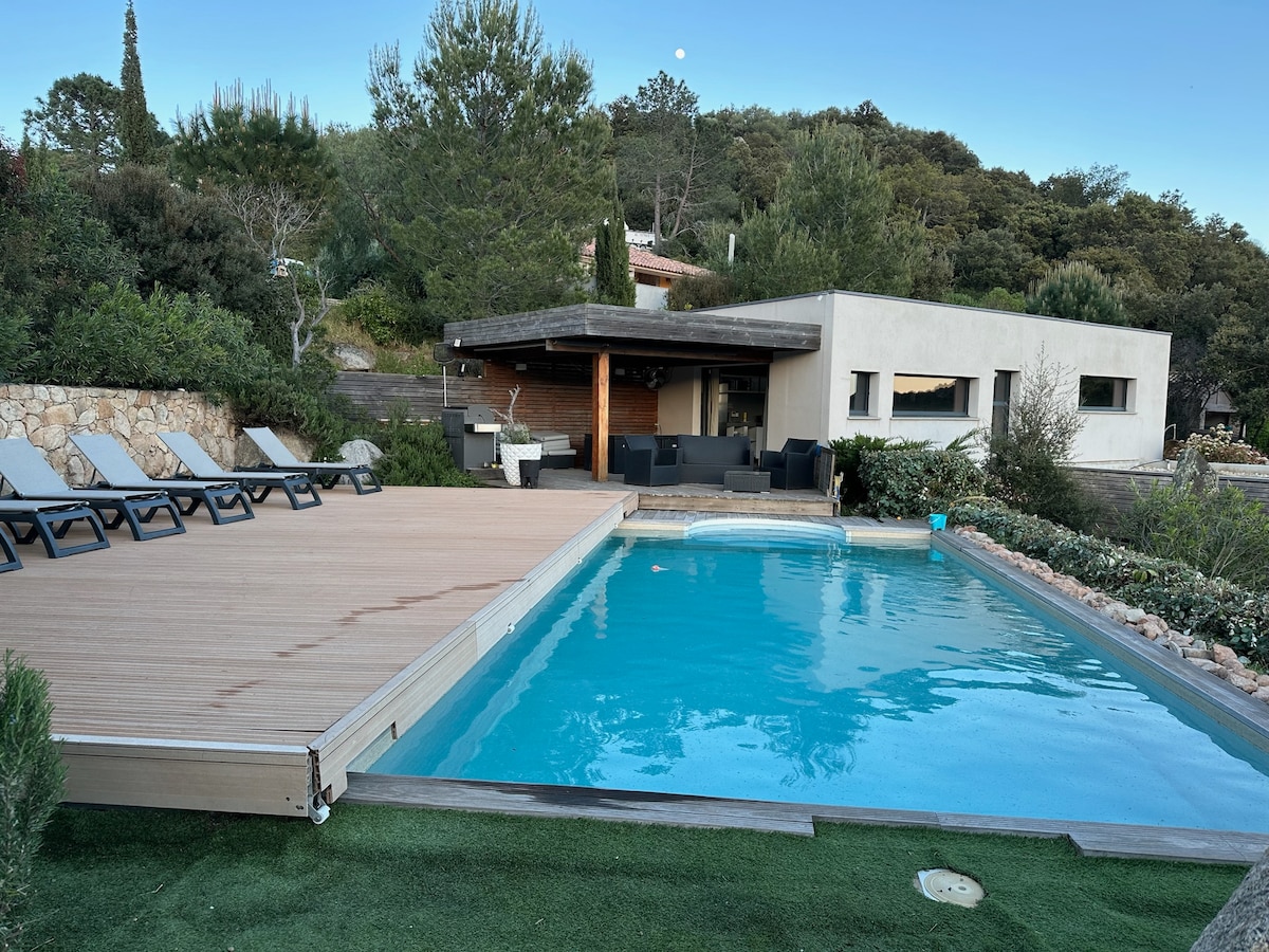 A serene outdoor area features a private salt-water pool with crystal-clear blue water. Surrounding the pool, a wooden deck is complemented by several lounge chairs. A modern villa is visible in the background, integrated into the lush landscape, with trees and greenery enhancing the setting.