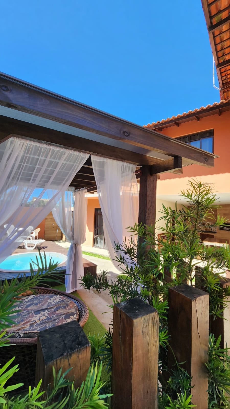 A pleasant outdoor area features a wooden gazebo draped with sheer white curtains, spanned over a comfortable seating arrangement. Lush greenery surrounds the gazebo, with a view of a private pool in the background, and a sunny sky overhead.