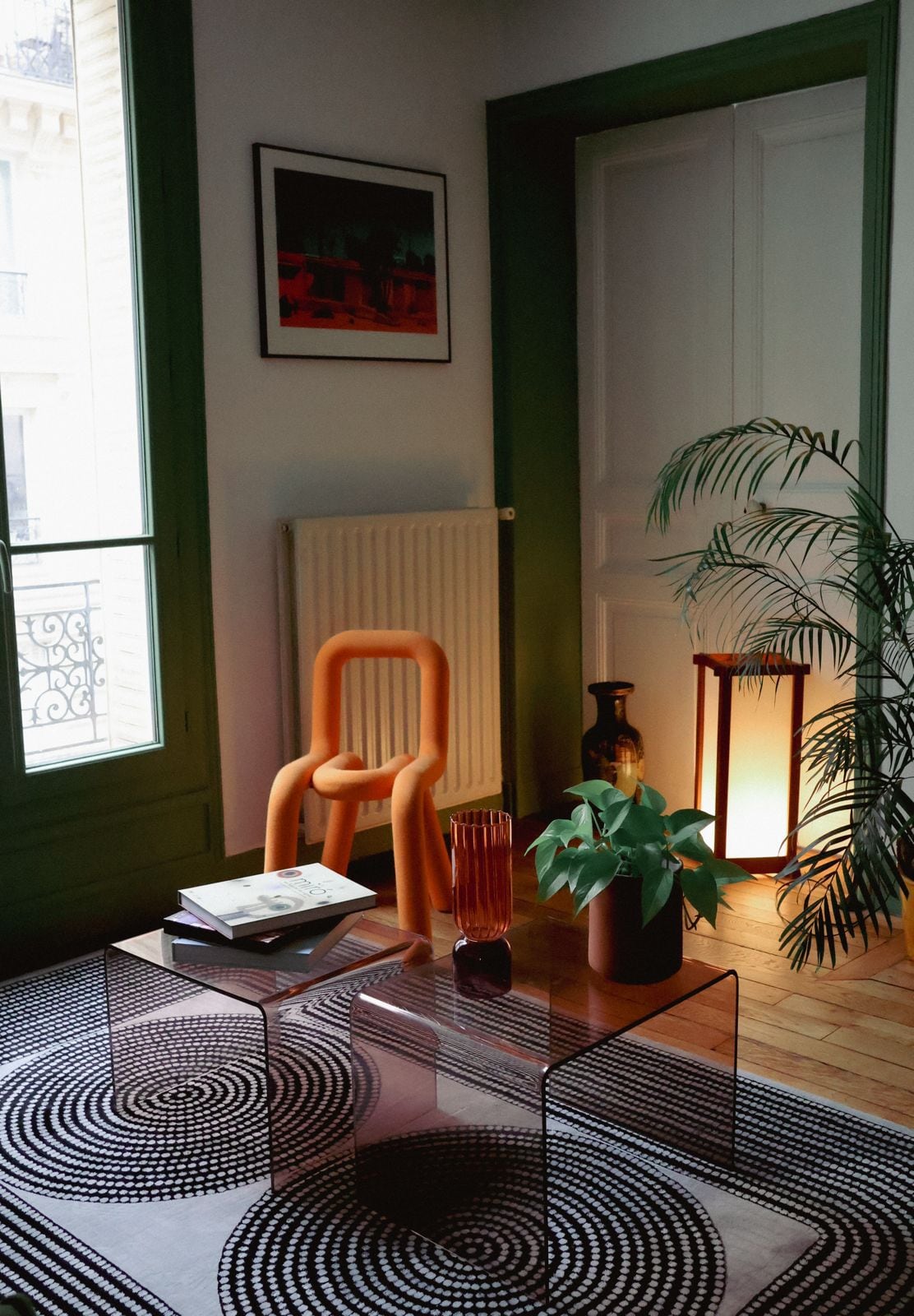 A stylish living space features a transparent coffee table and a modern orange chair. A potted plant and a decorative vase add greenery to the scene. Soft lighting from a floor lamp creates a warm ambiance, with a large window visible in the background.