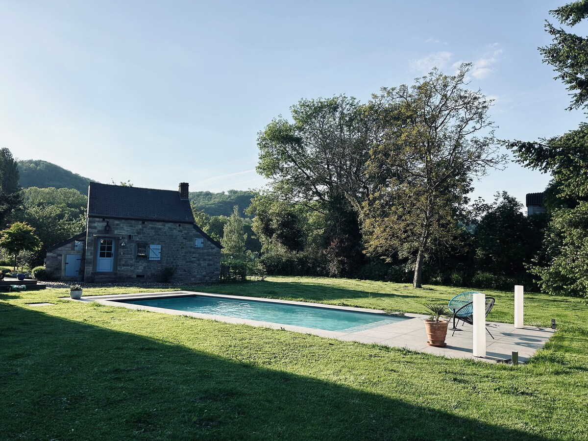 A serene outdoor space features a clear swimming pool surrounded by lush green grass. A charming stone house is visible in the background, framed by tall trees and distant hills, providing a tranquil setting for relaxation.
