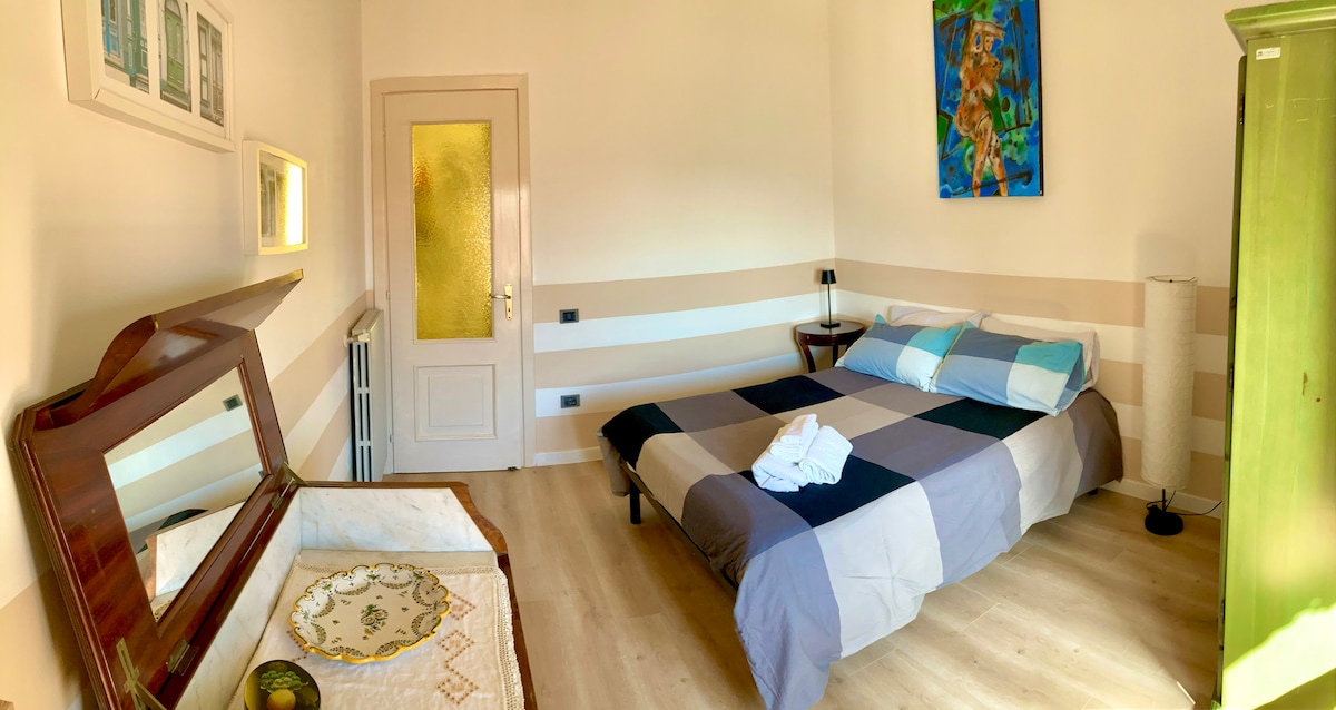 A neatly arranged room features a spacious bed with light blue and gray bedding, positioned against a light wall. An antique-style dresser is visible, alongside a decorative plate on a bedside table. Natural light streams in through a glass-paneled door.
