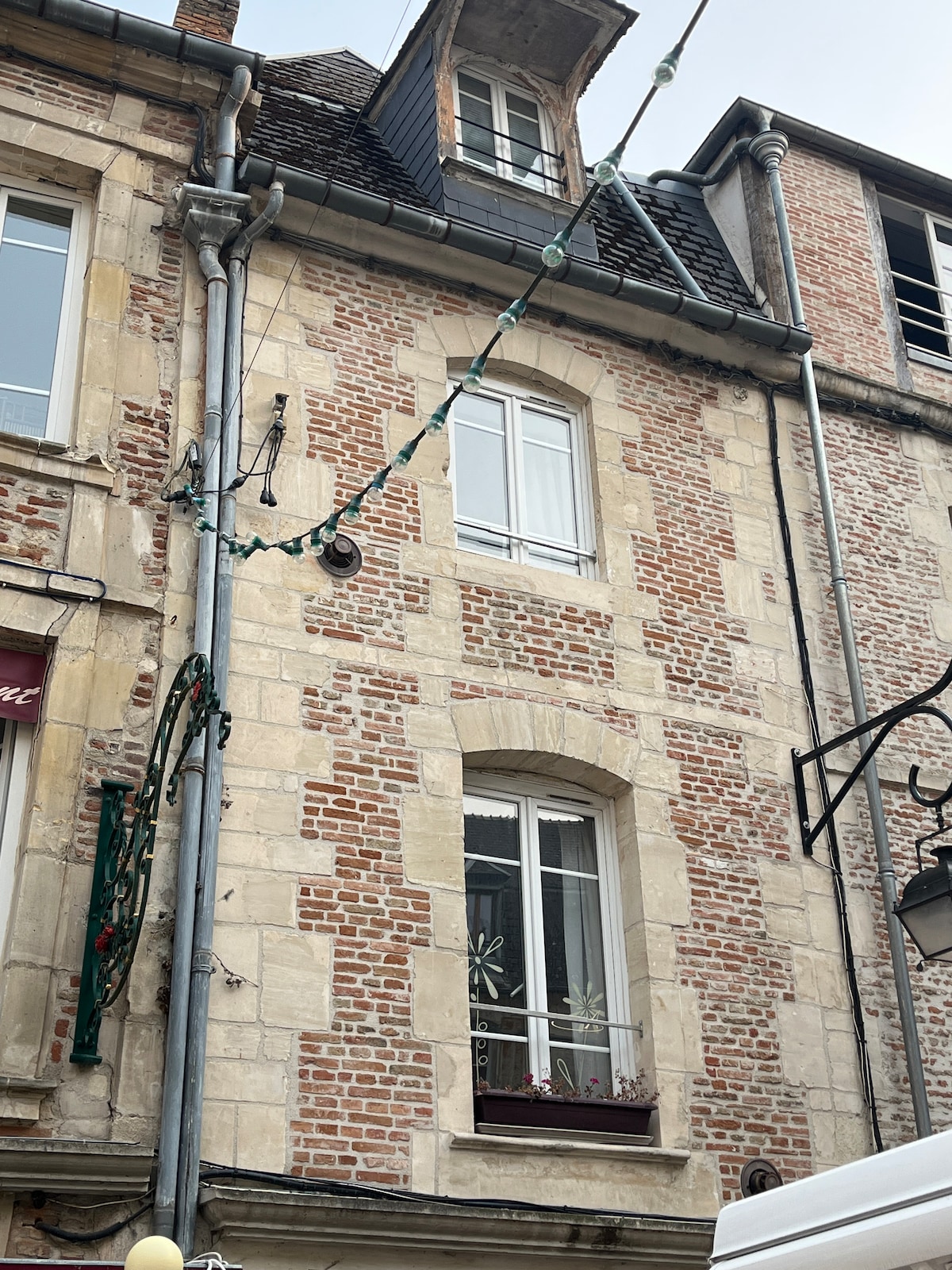 The image displays a charming brick building featuring a prominent window adorned with floral decorations. The textured façade is highlighted by a combination of light and dark bricks, with electrical wires and festive lights running above, creating a quaint urban atmosphere.