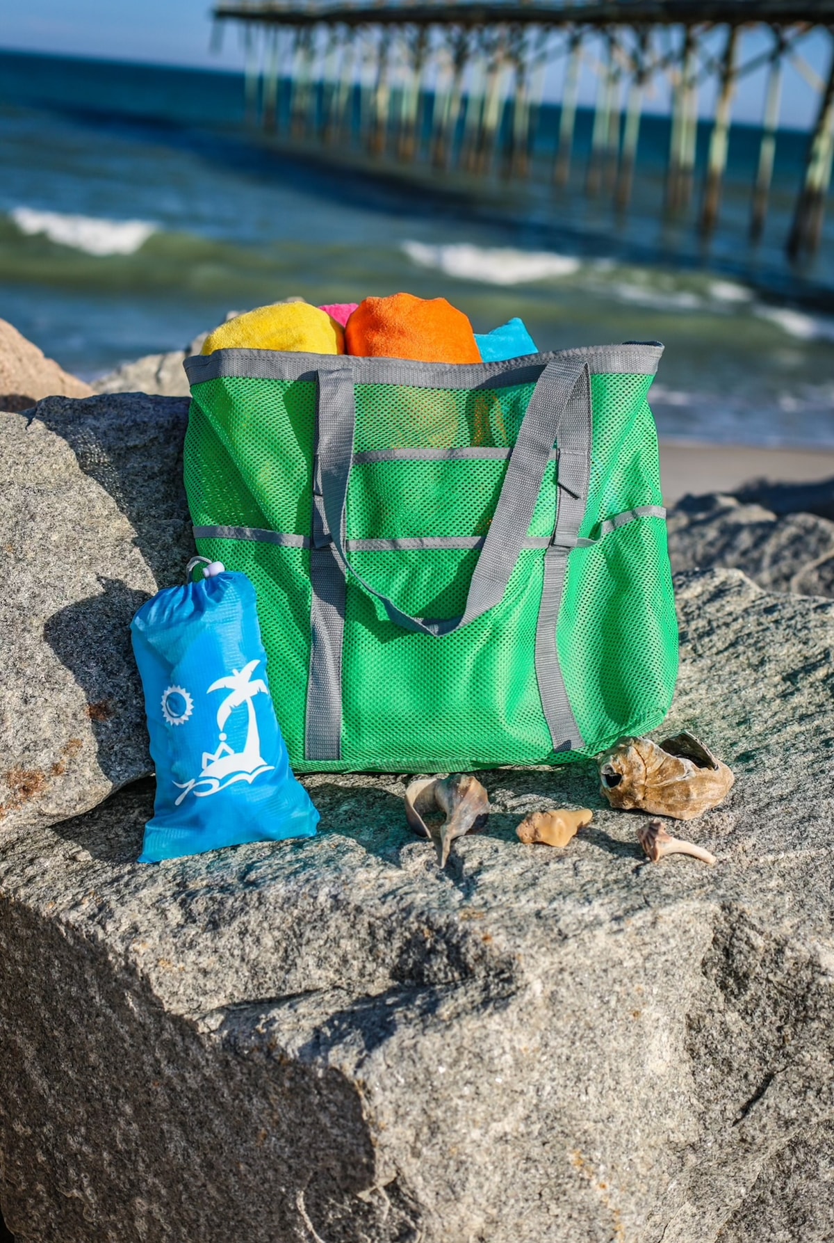 A green mesh beach bag is placed on a rocky surface, filled with colorful beach towels. Beside the bag, a blue pouch with a sun and palm tree design rests, along with several seashells. In the background, ocean waves gently lap against the shore under a clear sky.