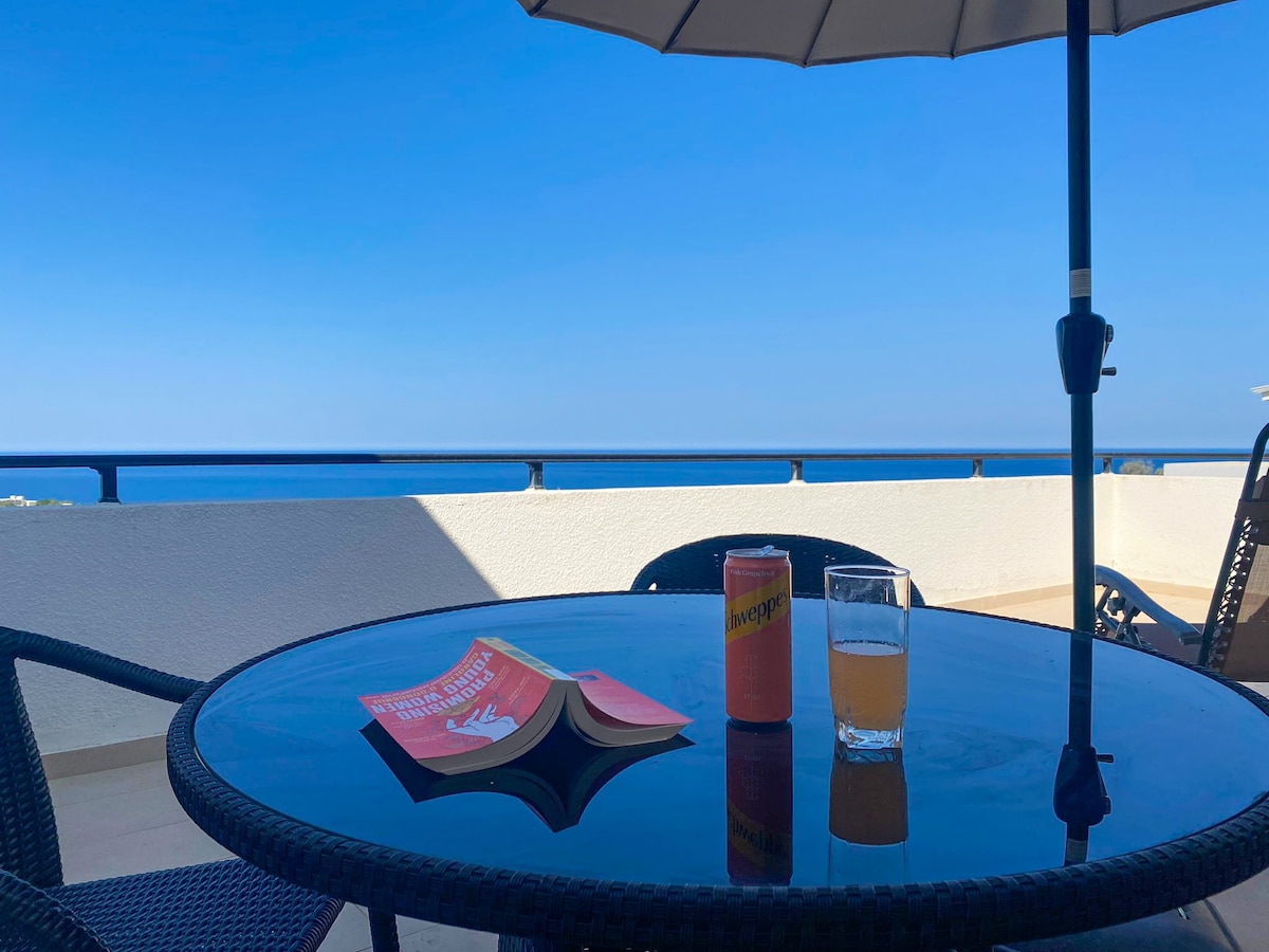 A terrace table with a glass and a can of drink sits under a shaded umbrella, offering a view of the Mediterranean Sea. A book is placed on the table, while clear blue skies create a serene backdrop.