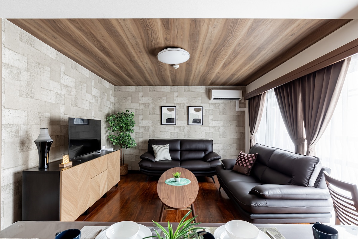 The living area features two dark leather sofas arranged around a round coffee table. A flat-screen TV is mounted on the wall opposite. Natural light filters through large windows adorned with curtains, highlighting the textured wall and wooden ceiling.