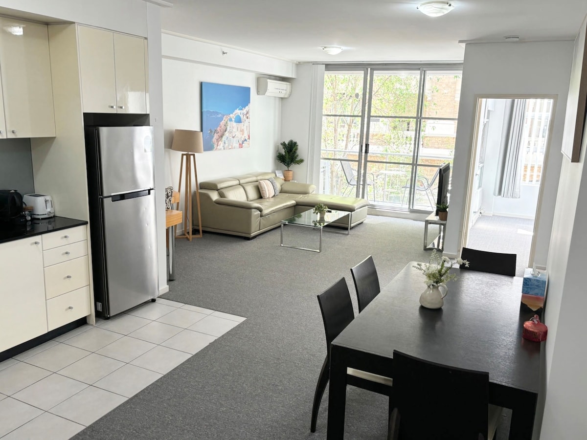 A modern open-plan living space features a sleek kitchen with stainless steel appliances and a dining table set for four. A light-colored sofa faces large windows that let in natural light, illuminating the cozy seating area and creating a welcoming environment.