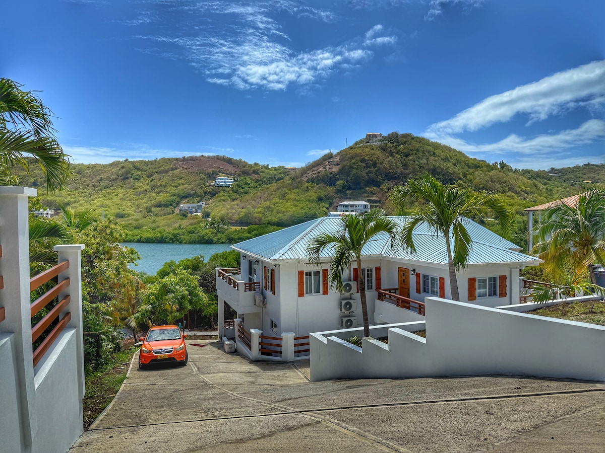 The exterior of the property displays a modern three-bedroom unit set in a lush green landscape. A driveway leads to the entrance, with a vibrant orange vehicle parked nearby. The backdrop features rolling hills under a clear blue sky.