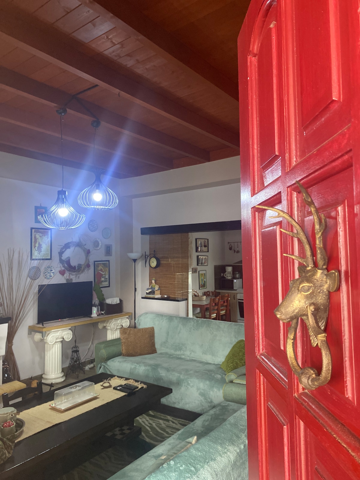 The view features a vibrant red door with a decorative handle, opening into a cozy living space. Soft lighting is provided by pendant lamps above, highlighting the green seating area and a dark coffee table. An inviting atmosphere is created with greenery and decorative accents throughout.