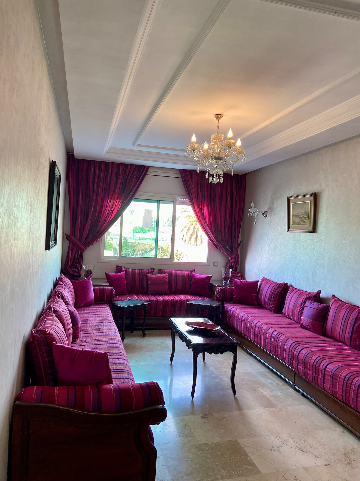 The living room features a harmonious arrangement of vibrant pink upholstery on spacious seating, complemented by a decorative chandelier overhead. Natural light filters through the window, illuminating the textured walls and marble flooring, creating an inviting space for relaxation and socializing.