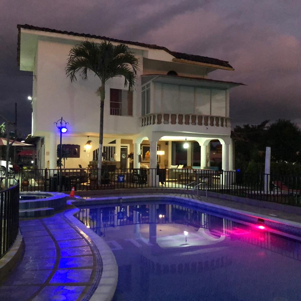 A spacious two-story building is depicted beside a well-lit pool area, featuring comfortable outdoor seating under a covered porch. The pool is illuminated with soft lighting, reflecting hues of pink and blue from underwater lights against a backdrop of a darkening sky.