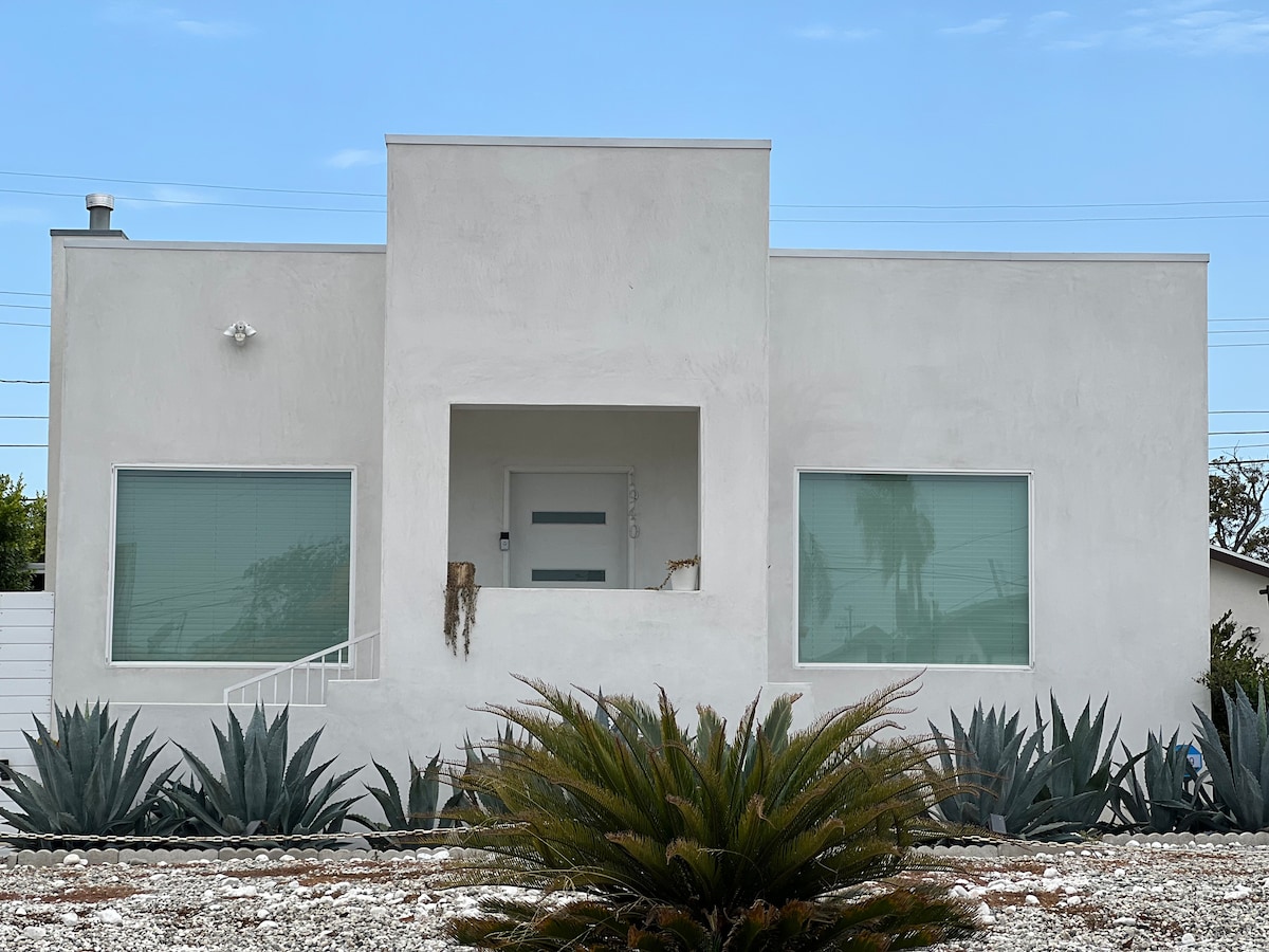 A modern, white stucco home features a centered entryway with a simple, sleek door. Large windows on either side allow ample natural light, while surrounding landscaping includes low-growing succulent plants and a textured rock garden for added visual interest.