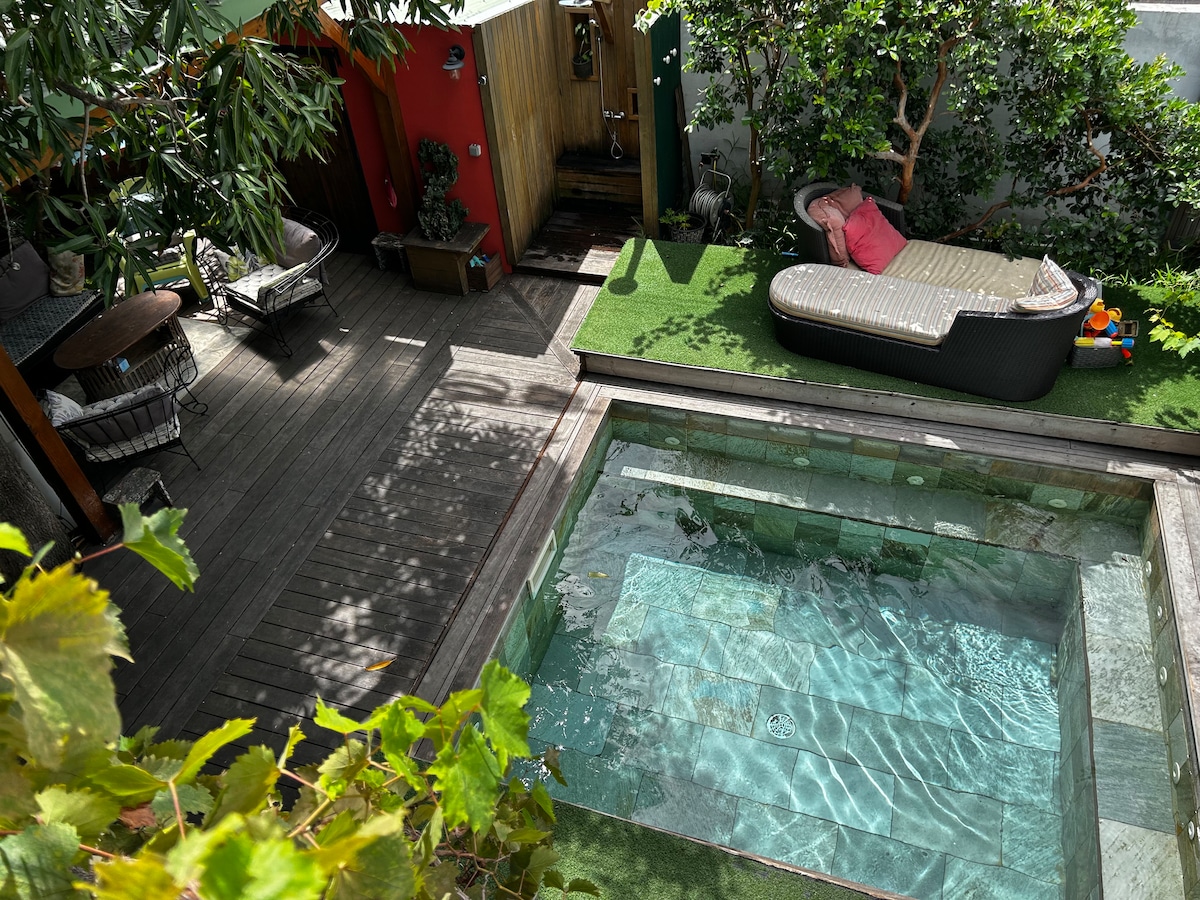 An outdoor area features a relaxation space with a comfortable lounge chair and two seating areas. A heated pool occupies the center, surrounded by wooden decking and vibrant greenery, creating an inviting space for family gatherings.