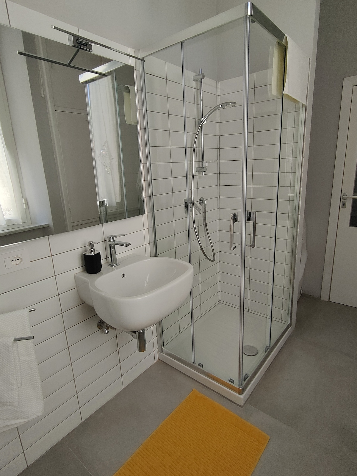A modern bathroom features a glass shower enclosure with a rainfall showerhead. A sleek, wall-mounted sink is positioned beside a large mirror. Neutral tiles complement a bright yellow bath mat on the floor, enhancing the clean and refreshing atmosphere.