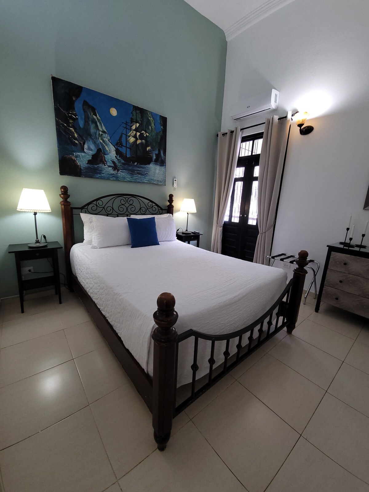 A room features a spacious bed framed by intricately carved wooden posts. Soft, neutral bedding complements the color palette of pale green walls. Two bedside tables with lamps provide soft lighting, while a large painting adds a maritime theme to the decor.