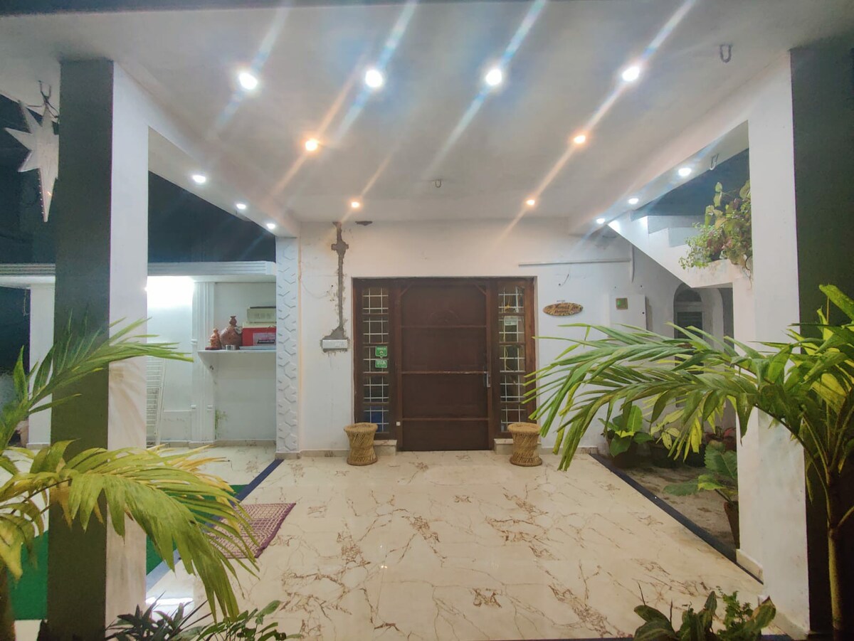 A spacious entrance area is illuminated by recessed lighting, highlighting the marble floor. Potted plants add greenery, and a large wooden door serves as the focal point. An open kitchen area is visible in the background, creating an inviting atmosphere for guests.