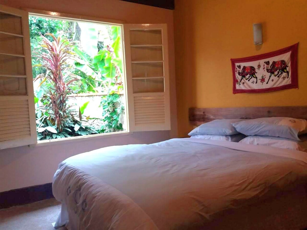 A bedroom features a spacious bed with neatly arranged white bedding. A large window offers views of lush greenery, enhancing the natural light that fills the room. Soft yellow walls create a warm and inviting atmosphere, complemented by decorative artwork on the wall.