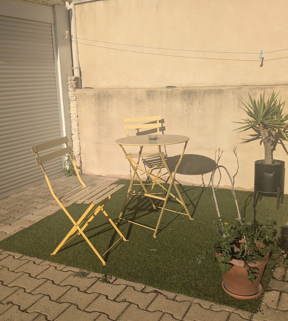 A small outdoor area features a light yellow bistro table with two matching folding chairs. Artificial grass covers the ground, providing a neat surface. A potted plant stands nearby, adding a touch of greenery, while a textured wall is visible in the background.