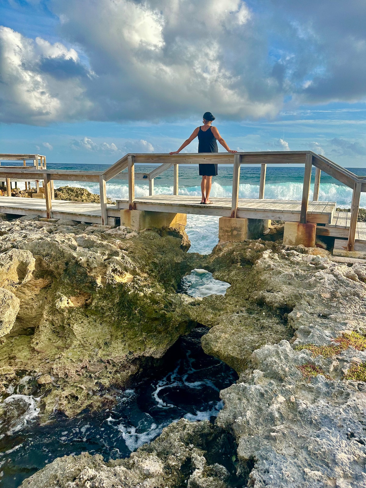 A wooden boardwalk extends over rocky shoreline, leading to crashing waves below. A person is seen standing at the end of the boardwalk, gazing out towards the sea. The sky is filled with soft, fluffy clouds, adding to the coastal ambiance.