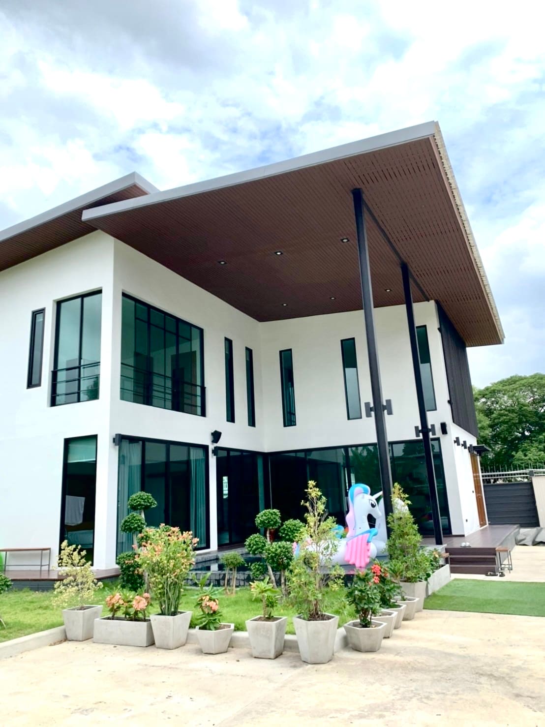 A modern two-story building features large glass windows and a striking overhanging roof. A landscaped garden, with neatly arranged potted plants, enhances the inviting exterior. A whimsical unicorn statue adds a playful touch to the outdoor area.