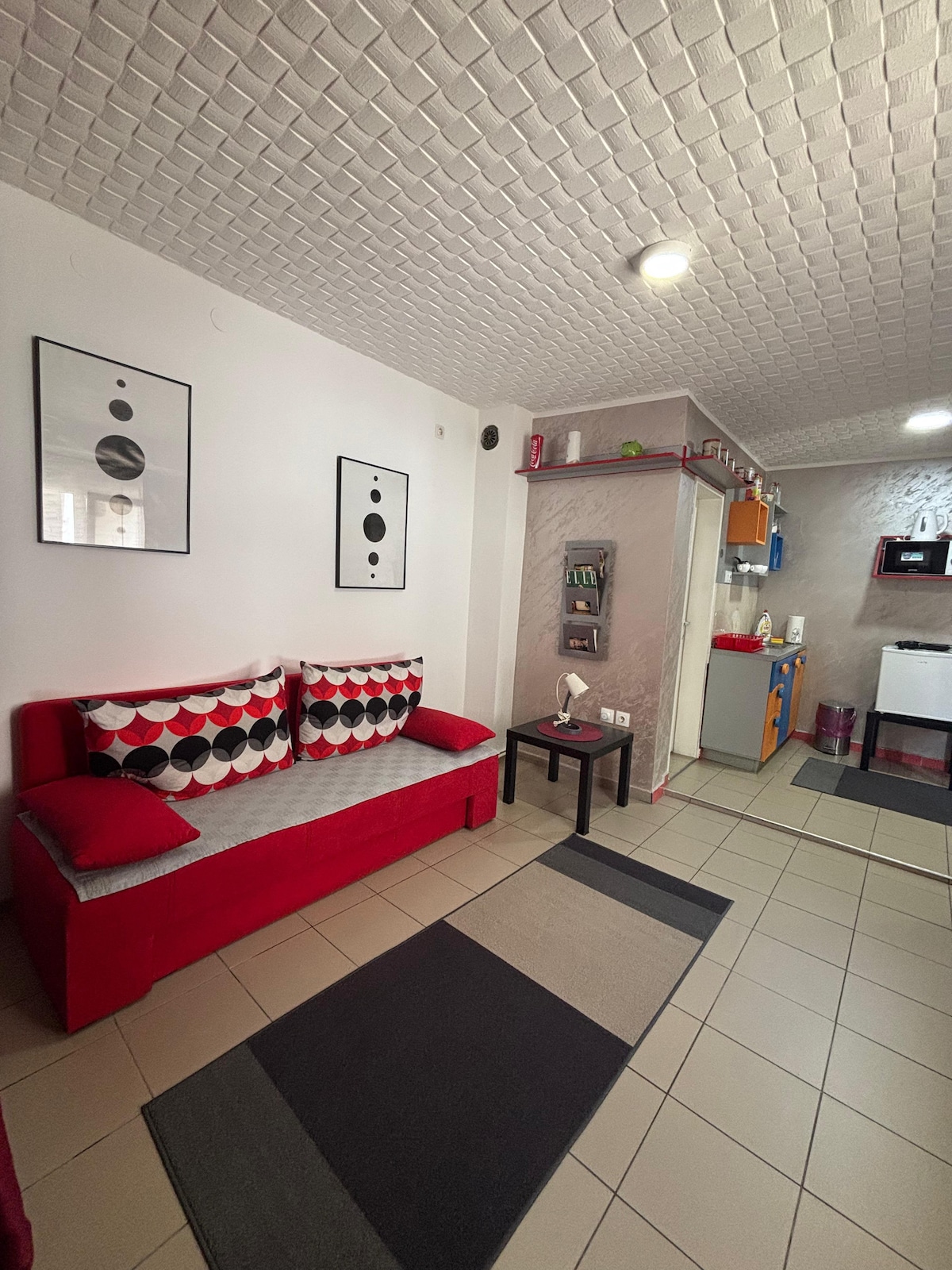 A compact living area is presented, featuring a bright red sofa adorned with patterned cushions. A neutral-toned rug lies beneath, complementing the tiled floor. A kitchenette is visible in the background, equipped with cabinets, a microwave, and a small refrigerator, providing essential amenities.